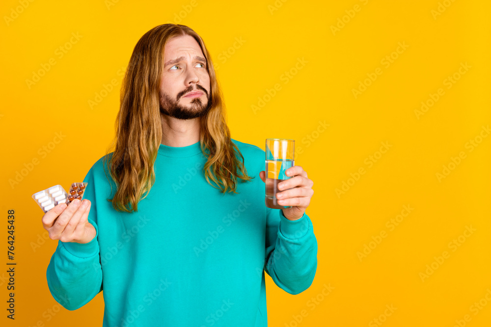 Portrait of unsatisfied minded guy hold pills glass water look empty space isolated on yellow color background Portrait of unsatisfied minded guy hold pills glass water look empty space isolated on yellow color background