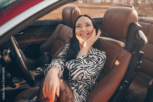 Happy smiling brunette woman driver sitting in new red cabriolet car on beach coast, smiling looking at camera enjoying journey. Driving courses and life insurance concept. Glamorous lifestyle