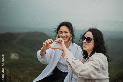 Young beautiful happy lesbian couple posing in cloudy mountains showing love sign outside at nature summer park. LGBT community concept. Female friends smiling enjoying moments together.