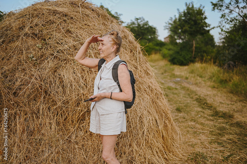Elderly senior traveling backpacker mature woman tourist smiling posing with stack of hay in field outdoors in countryside. Retired people summer holiday vacation, active lifestyle concept