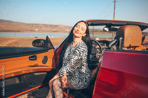 Happy smiling brunette woman driver sitting in new red cabriolet car on beach coast, drinking glass of champaigne. Concept of birthday celebration and luxury living. Glamorous lifestyle