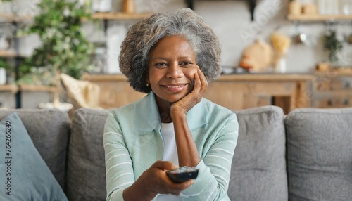 Bored senior woman spends leisure time watching TV shows, movies. The mature elderly female sitting on the couch alone with a remote controller and switching channels, feels drearily. Front view 