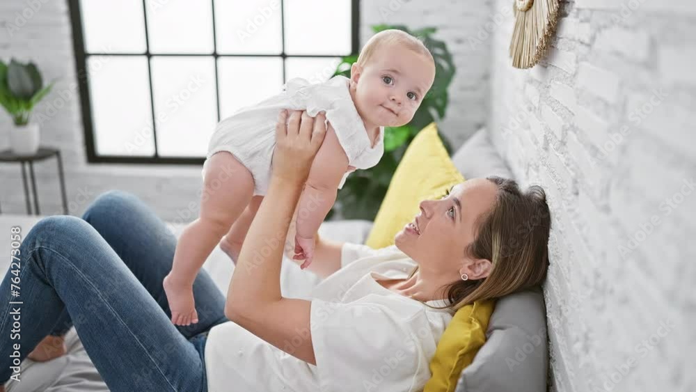 Joyful mother lying on bed, confidently holding happy baby daughter mid-air, having fun while smiling and playing together in warm, homely bedroom. Joyful mother lying on bed, confidently holding happy baby daughter mid-air, having fun while smiling and playing together in warm, homely bedroom.
