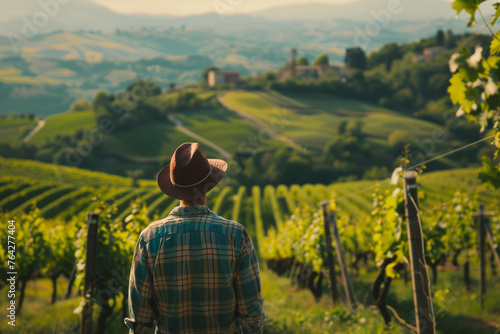 A man wearing a hat stands in a vineyard. The vineyard is lush and green, with rows of vines stretching out in front of him. The man is taking in the beauty of the landscape
