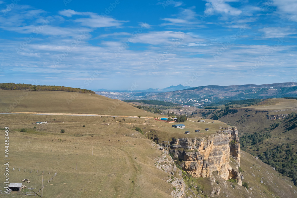 Foothills of the Greater Caucasus. Panoramic aerial view of a picturesque gorge with a farm on a rocky cliff. A populated area in the distance.