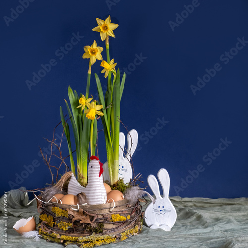 Easter decor in rustic style. Nest, chicken, two funny hares and yellow daffodils on dark blue background. Handmade. Concept of home comfort and decor for bright holiday of Easter. Square.