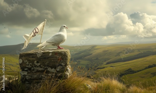 White pigeon standing on a stone pedestal with a peace flag waving in the breeze behind it against a backdrop of rolling hills