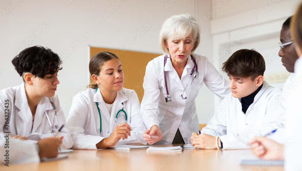 Obraz premium Group of medical workers in white coats sitting around desk during professional training course, listening to experienced aged female lecturer