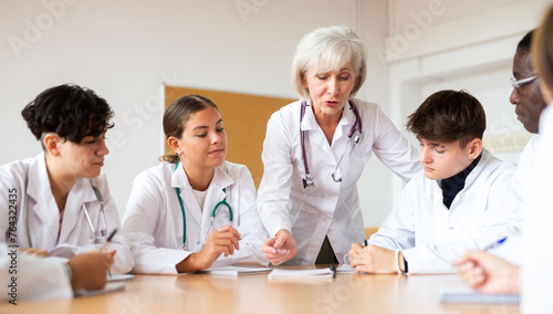Group of medical workers in white coats sitting around desk during professional training course, listening to experienced aged female lecturer