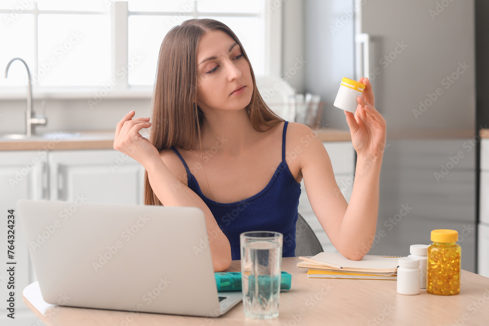 Beautiful woman with bottle of vitamin supplements at table in kitchen Beautiful woman with bottle of vitamin supplements at table in kitchen