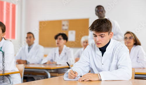Interested young student in white coat listening to lecture and taking notes in classroom during professional medical training