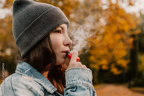 Young woman outside autumn park fall and smoking tobacco device electronic cigarette heater. Smoke and steam system with sticks inside, image with copy space. Harmful habit harm to health lungs 