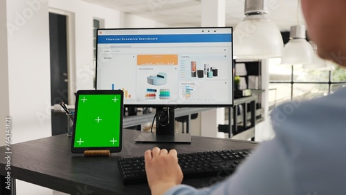Employee checks tablet with greenscreen while she works on business operations at desk, looking at modern gadget presenting isolated copyspace display. Woman looks at mockup layout. Handheld shot.