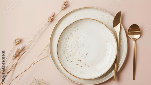 A tasteful beige empty porcelain plate with gold cutlery on a soft pastel pink background  adorned with delicate dried flowers.