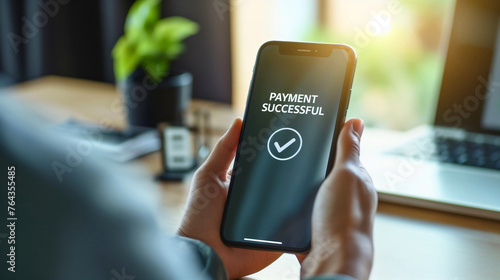 Businesswoman holding phone with text payment successful on screen display, sitting in office chair at desk or table with laptop notebook on workplace. Online internet shopping, purchase invoice