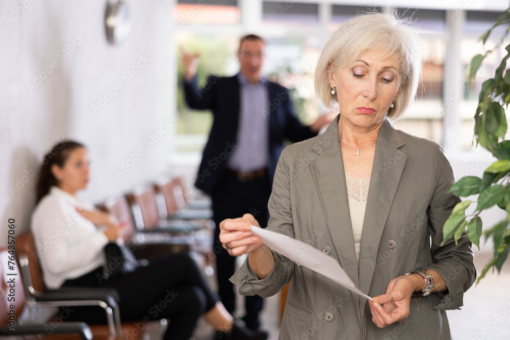 Upset old woman holding white paper, standing in waiting room with her back to person who is shouting at her