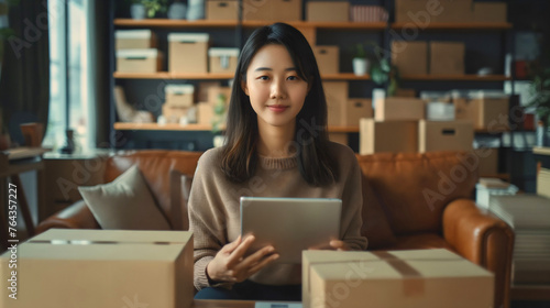 Beautiful young Asian businesswoman holding tablet device, smiling and sitting surrounded by cardboard boxes ready for shipping. Delivery package distribution, online shop stock, commerce inventory © Nemanja