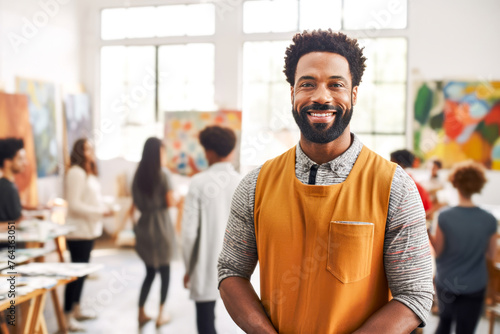 African American male smiling art teacher in mustard apron stands in art studio. Group of focused students on background. Concept of nurturing creativity and mentorship in art class education