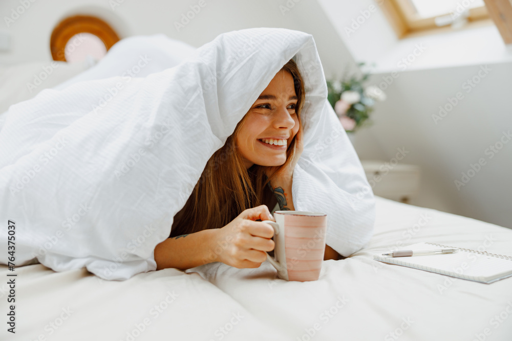 A happy woman wrapped in a blanket is laying in bed and holding a cup of coffee with smiling A happy woman wrapped in a blanket is laying in bed and holding a cup of coffee with smiling
