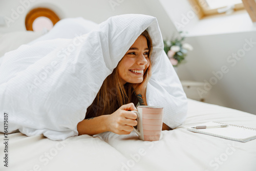 A happy woman wrapped in a blanket is laying in bed and holding a cup of coffee with smiling