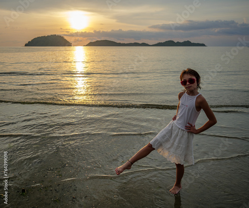 young girl in a white dress on the beach against the backdrop of sunset (ID: 764399098)