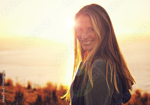 Sunset, happy and portrait of woman by ocean for adventure, relaxing and enjoy weekend outdoors. Nature, smile and person with flare on hill for holiday, vacation and travel destination in evening © Mariusz S/peopleimages.com