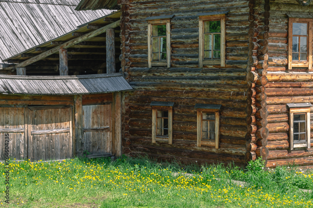 A two-storey old house in the village made of dark logs with a wooden roof. Yellow dandelions in front of the house in the countryside. Wooden old manor house of the 19th century. A two-storey old house in the village made of dark logs with a wooden roof. Yellow dandelions in front of the house in the countryside. Wooden old manor house of the 19th century.