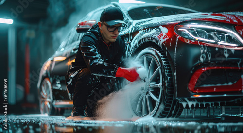 A man in black and red , wearing glasses with a cap is washing the wheels of his shiny car in white foam on a dark background. He looks focused while doing it
