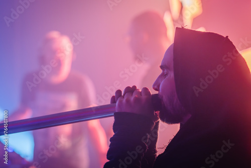 Valmiera, Latvia - March 15, 2024 - A man in a hooded robe plays the didgeridoo, with others in soft focus behind him under pink and blue stage lights. photo