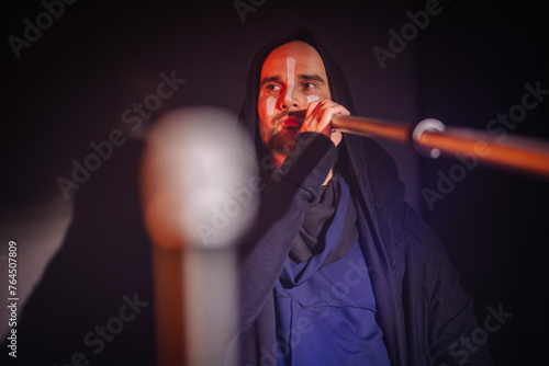 Valmiera, Latvia - March 15, 2024 - A man with face paint plays a didgeridoo, focused and serious, under dramatic side lighting with a microphone in the foreground...