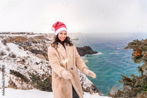 Outdoor winter portrait of happy smiling woman, light faux fur coat holding heart sparkler, posing against sea and snow background © svetograph