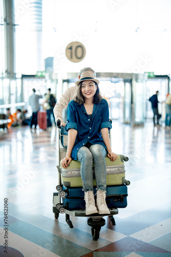 Portrait of happy Asian young girl sitting on luggage by Grandpa pushes a cart in international airport.