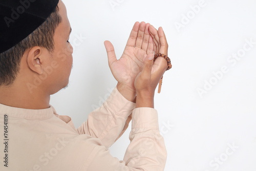 Back view portrait of religious Asian muslim man in koko shirt with skullcap praying earnestly with his hands raised, holding islamic beads. Devout faith concept. Isolated image on white background