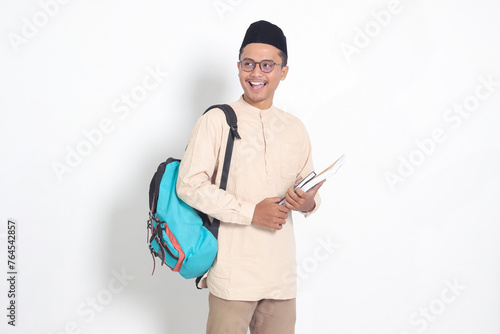 Portrait of excited student Asian muslim man in koko shirt with skullcap carrying backpack, while holding his school books. Islamic education concept. Isolated image on white background photo