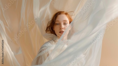 Beautiful redhead young woman wrapped in transparent fabric. Fluttering veil on a beige background