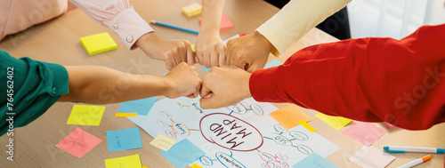 Young happy creative startup group join circle fist bump hands together surrounded by marketing strategy mind map and colorful sticky notes at meeting room. Unity and teamwork concept. Variegated. photo