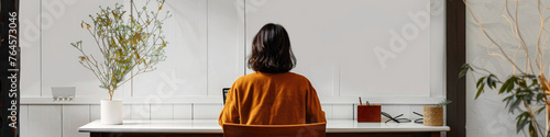 A woman sits at a white table facing away, concentrating on a laptop screen, with a plant and soothing interior decor