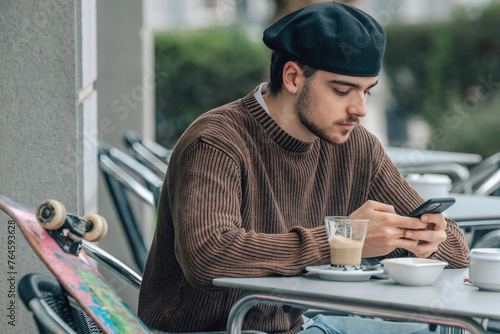 young urban man on the cafe terrace with mobile phone and skateboard