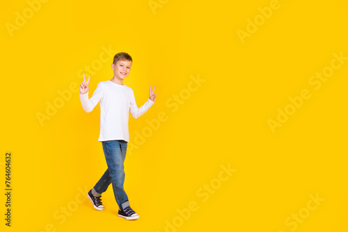 Full length photo of cheerful positive boy wear white shirt walking showing v-signs empty space isolated yellow color background