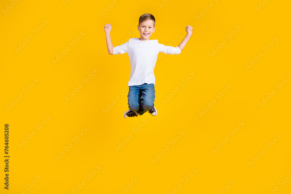 Full length photo of carefree lucky boy wear white shirt jumping high rising fists isolated yellow color background