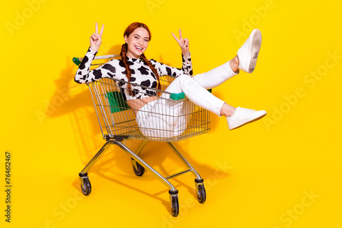 Full body photo of charming friendly lady sit inside hypermarket trolley show v-sign isolated on yellow color background