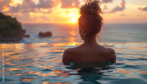 back view young adult female person enjoy relax in infinity edge luxury outdoor swimming pool looking on ocean view in warm sunset light. Tropical wellness travel vacation tourism concept