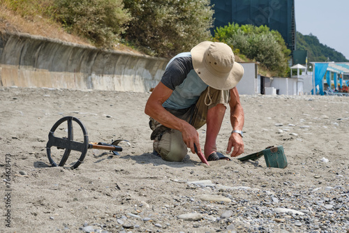 A man wearing shorts and a hat, with a metal detector, is searching for gold and coins on a sandy beach the daytime in summer.