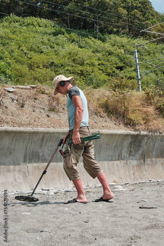 A man wearing shorts and a hat, with a metal detector, is searching for gold and coins on a sandy beach the daytime in summer.