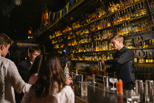 Pub guests supervise bartender making cocktails sitting by counter. Barkeeper with shaker in hands background stylized shelves with alcohol