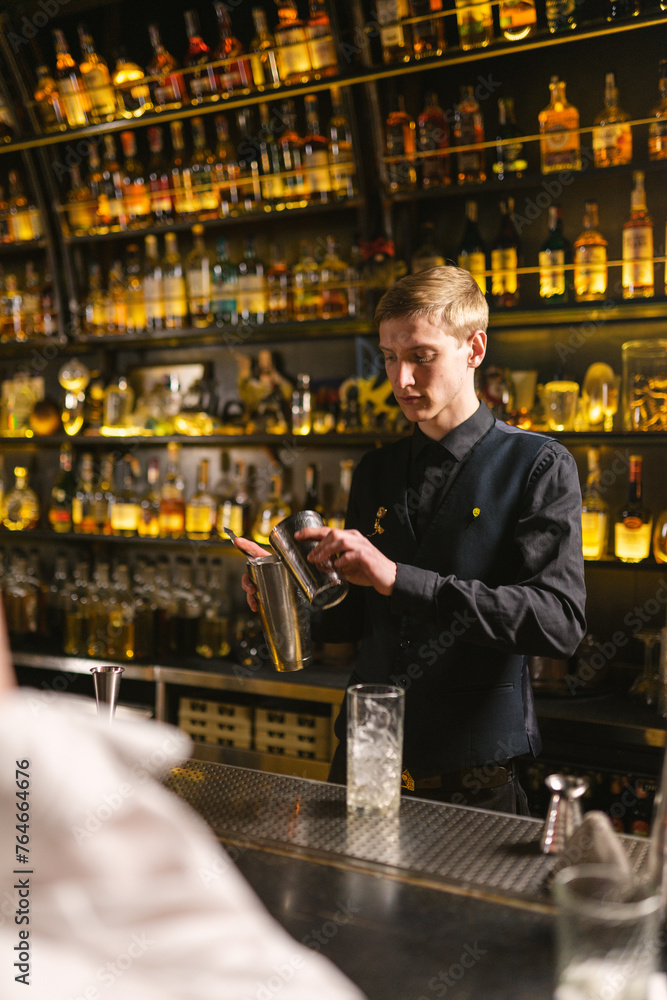 Focused bartender in black uniform works at bar counter making cocktails with aluminum utensils. Preparation of alcoholic drinks mixing in shaker for pub visitors