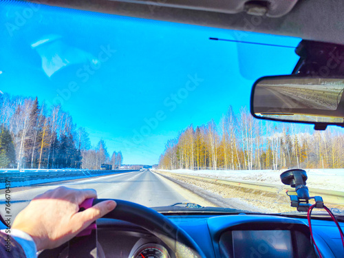 Winter Driving on winter or spring Snow Lined Road Under sunny blue Sky. Drivers perspective on snowy road with sun And hand of woman on steering wheel