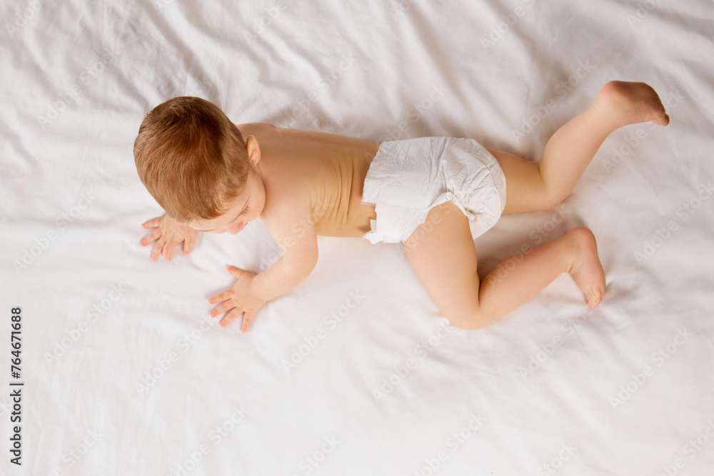Cute little baby boy, child in diaper lying on bed, crawling, playing isolated over white studio background. Concept of childhood and family, care, parenthood, infancy and heath Cute little baby boy, child in diaper lying on bed, crawling, playing isolated over white studio background. Concept of childhood and family, care, parenthood, infancy and heath