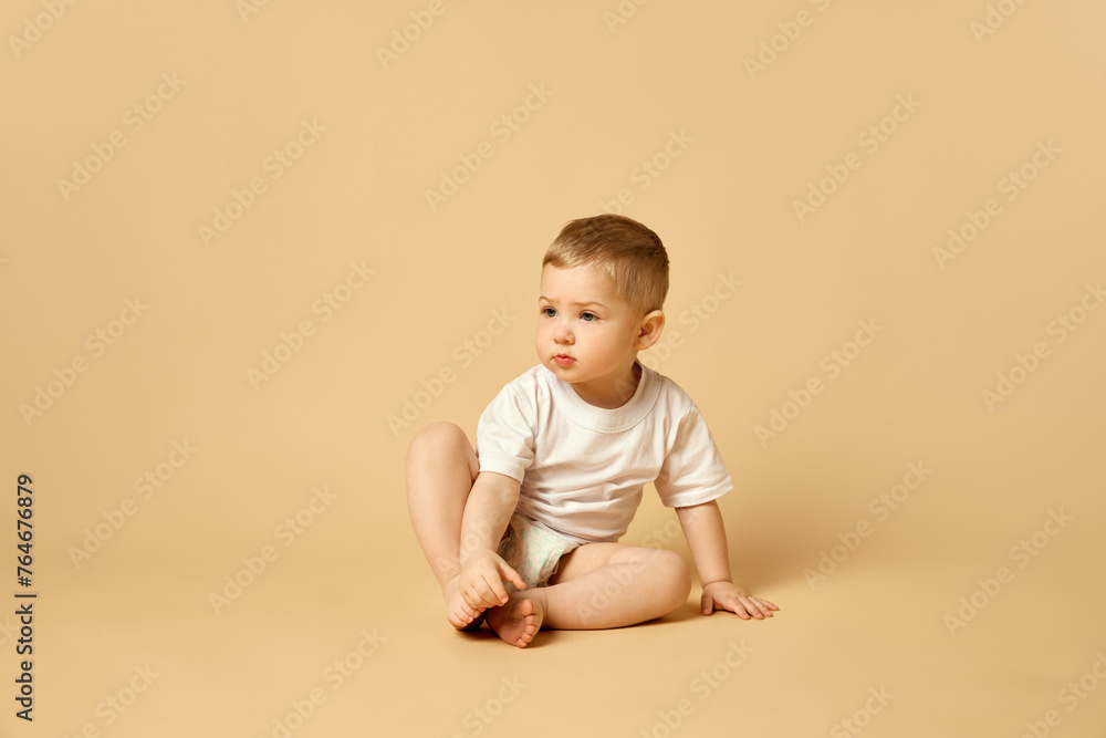 Cute little baby, boy, child in diaper and t-shirt calmly sitting on bed isolated over beige studio background. Concept of childhood and family, care, parenthood, infancy and heath Cute little baby, boy, child in diaper and t-shirt calmly sitting on bed isolated over beige studio background. Concept of childhood and family, care, parenthood, infancy and heath