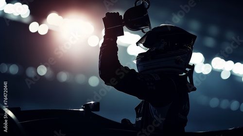 Silhouette of a racing car driver with helmet on, holding a trophy aloft in a victorious gesture against a backdrop of bright lights.